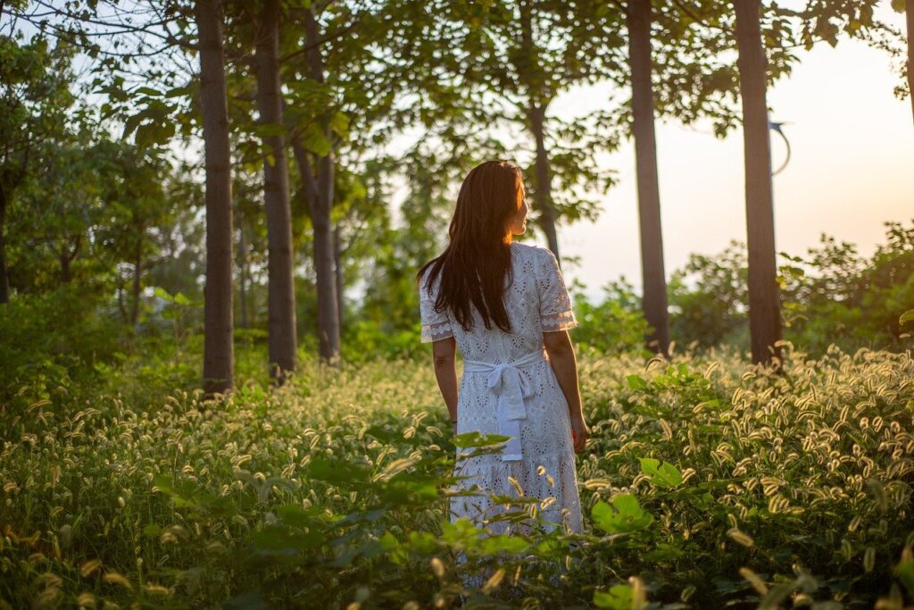 A woman outdoors in nature glancing at the sun glare. She is standing in the field of green grass with lots of tall trees nearby and wearing a white dress